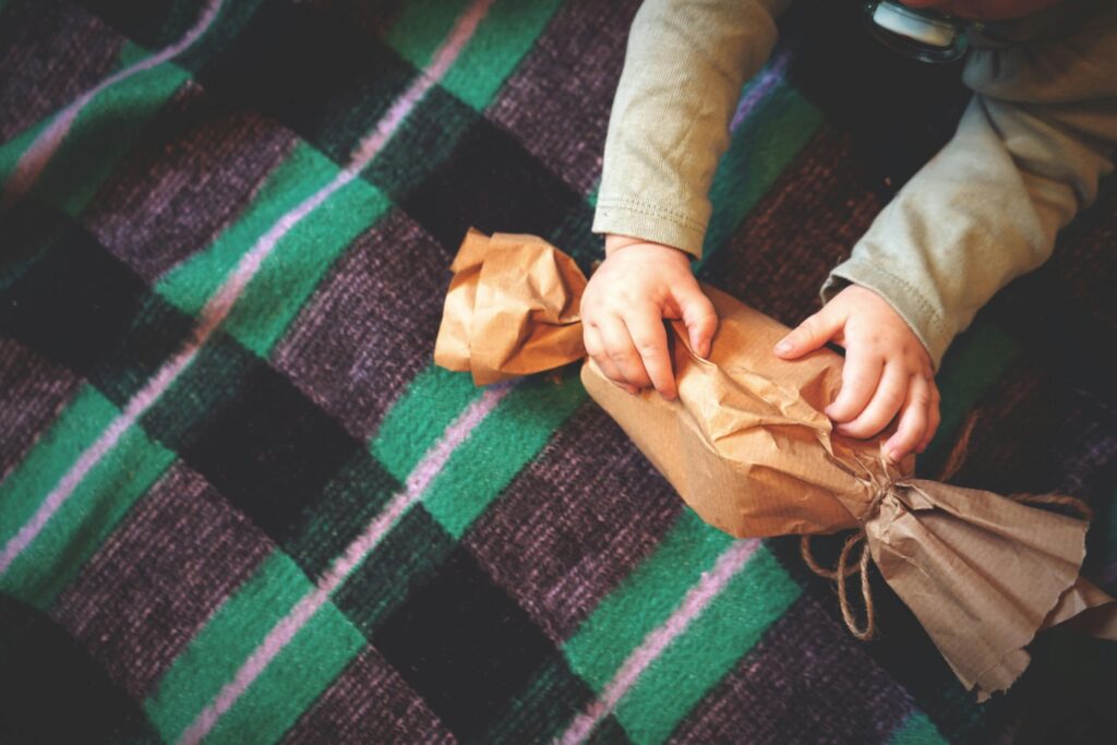 A child unwrapping a brown paper gift on a colorful checkered blanket.